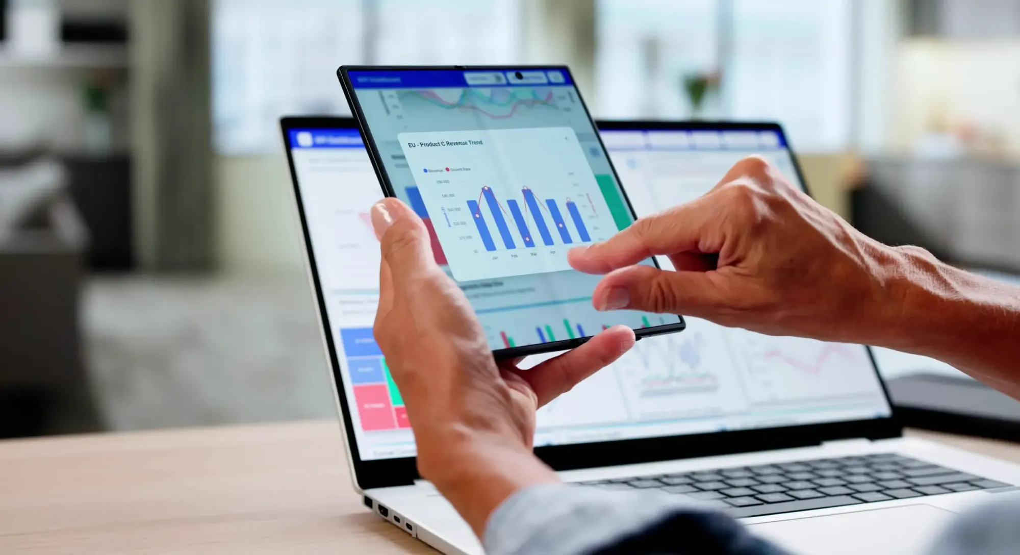 Person holding a tablet displaying a bar chart dashboard in front of a laptop with analytics data in a modern office.