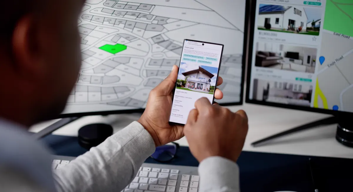 Real estate agent reviewing property listings on a phone and dual computer monitors.
